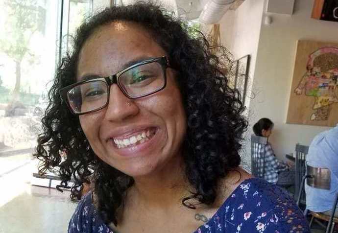 Biracial woman with curly black hair is smiling for the camera. She has black-rimmed glasses and is wearing a blue, flowered romper. She is seated in a coffee shop, backs of patrons visible behind her.