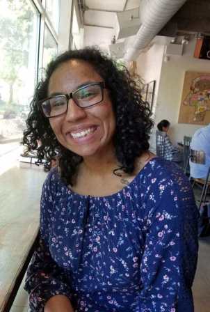 Biracial woman with curly black hair is smiling for the camera. She has black-rimmed glasses and is wearing a blue, flowered romper. She is seated in a coffee shop, backs of patrons visible behind her.