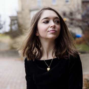 Woman with light white skin and long, dark brown hair is in the center of the image. She is looking toward the right, slightly smiling. She is wearing a black long sleeve shirt and behind her are trees and water.