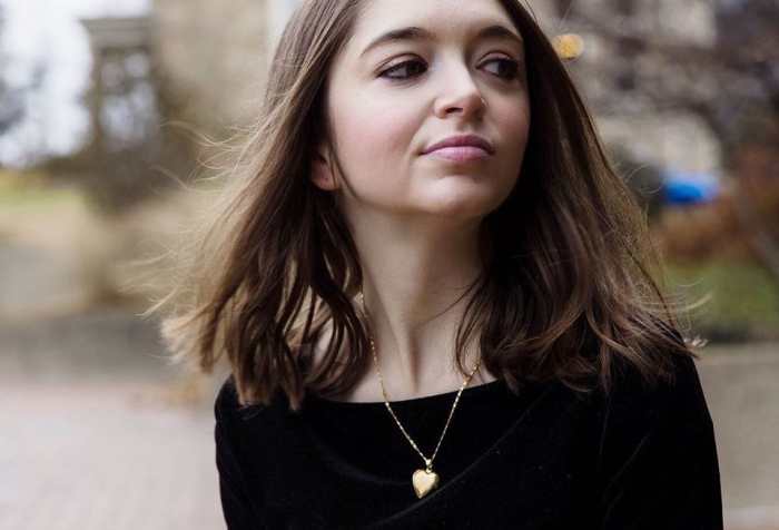 Woman with light white skin and long, dark brown hair is in the center of the image. She is looking toward the right, slightly smiling. She is wearing a black long sleeve shirt and behind her are trees and water.