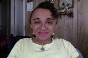 Young Black woman with her hair parted to the side and curled. She is smiling at the camera and wearing a yellow t-shirt. Behind her is room w/ wood panelling and a calendar and bird figurine on the wall.