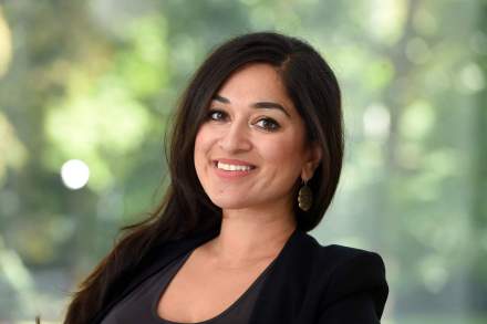 Jaipreet Virdi, a South Asian woman with dark hair, smiles at the camera. Her head is slightly turned. She is wearing a black blazer and dark top. There are some trees in the blurred background.