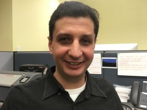 Photo of Walei Sabry smiling, wearing a black collared shirt, inside an office cubicle.