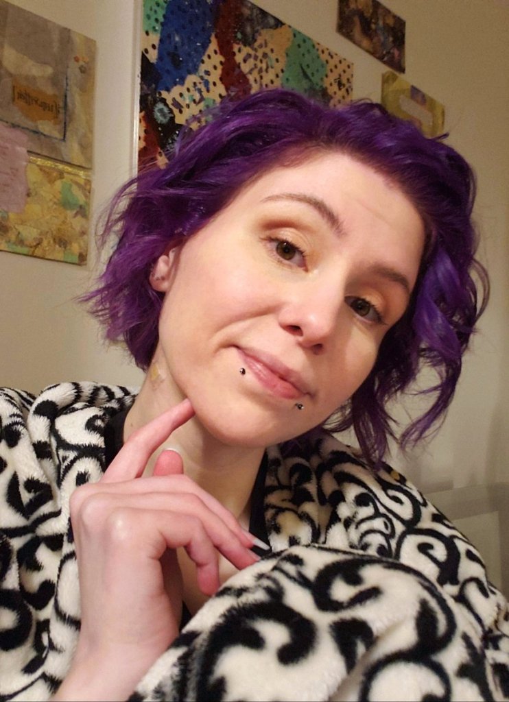 Kaalyn is sitting looking to the camera with a light smile and forefinger to chin. She has purple chin-length hair, loosely curled, minimal makeup, and two black snakebite piercings at the lip. She's wrapped in a black and white decorative blanket in front of a wall of very colorful abstract art.