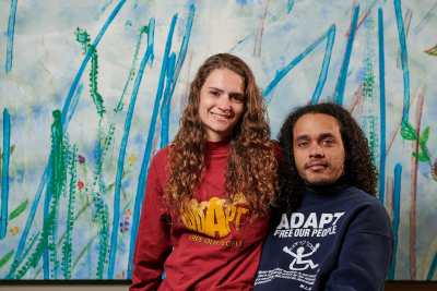 Photo of life-partners and Co-Executive Directors of The Partnership for Inclusive Disaster Strategies, Shaylin Sluzalis and Germán Parodi. On the left is Shaylin Sluzalis, a white woman with long brown curly hair, in a red long sleeve ADAPT shirt smiles as she sits on the side of Germán Parodi's wheelchair who is on the right of Shaylin. Germán Parodi, a Puerto Rican man is sitting in his manual wheelchair with long dark brown curly hair, in a long navy blue ADAPT shirt with a slight grin and his arm around Shaylin. Their background is a light blue painting with multi-colored lines from wheelchair wheels with paint, a project done by disabled Philadelphian's with Disability Pride Philadelphia. Photo credit: Albert Yee