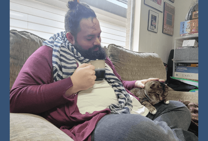 Graphic with a blue-gray background. In the center is a photo of a white, nonbinary, disabled fat person sits on a tan couch with their legs folded. They are wearing a wine red cardigan and a blue striped scarf, and have long, dark curly pulled back in a hair clip and have a dark beard. They are drinking a cup of tea and looking down at a tan cat sitting in their lap and petting the cat.