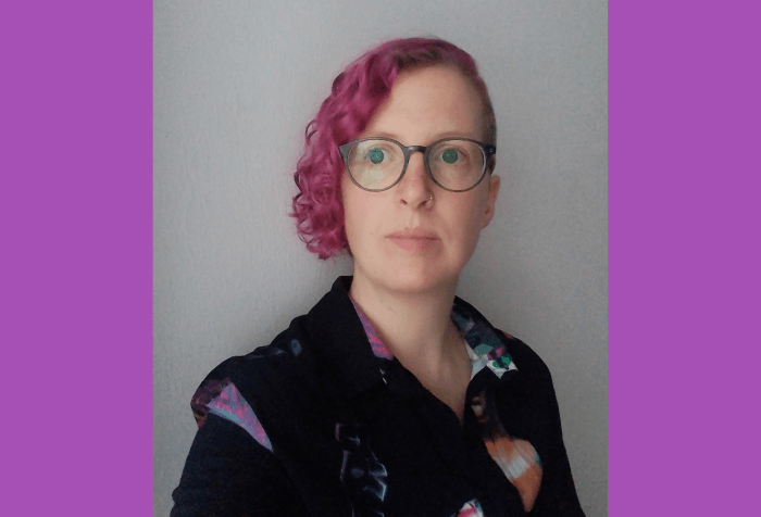 Graphic with a purple background. In the center is a photo of a white woman against a grey wall, her head is tilted slightly to the right, looking away from the camera. She has curly hair, dyed magenta, uses greenish glasses and is wearing a black shirt.