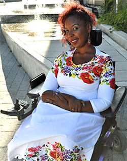 Image description: Andraéa sitting in her power wheelchair wearing a long, white dress with brightly colored flowers lining the collar and bottom of the dress. She is sitting in front of a wooden ramp with water and the sun shining brightly behind her.