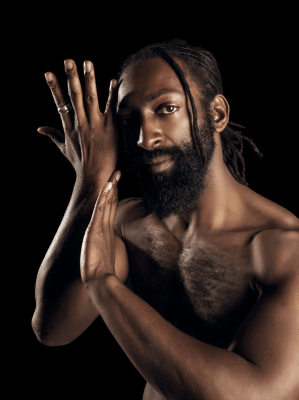 image of Antoine Hunter, a self-described handsome, dark milk chocolate, African American Man. He has long Ebony dreadlocks hair tied to back leaving one deadlock on the side of his face which is close to his eyes. He has a full 1-inch long Ebony-colored bread and full brown lips. He is bare chested and his right hand is to the side of his face with his left hand touching his right forearm. His eyes look straight at you with honest spirit. 