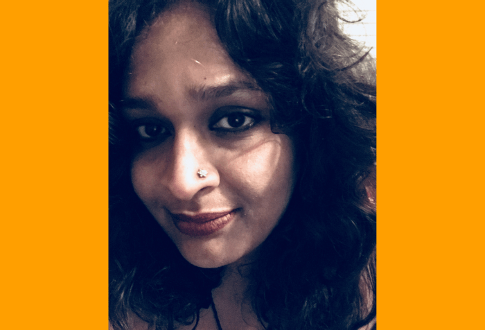 Orange background with a color photo showing a selfie of an unsmiling Indian woman, with curly dark brown hair and dark eyes. She has a nose stud on her left nostril.