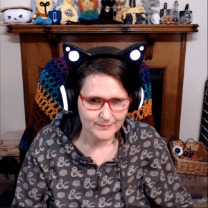 Young white woman wearing pink-framed eyeglasses and a hoodie. She is also wearing headphones that look like black cat ears. Behind her is a wooden mantle full of cute nerdy stuffed creatures.