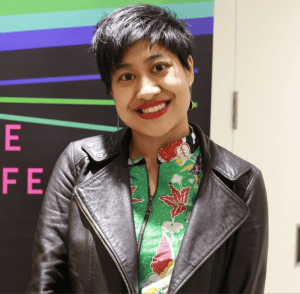 Image description: Khairani Barokka, a young Indonesian woman with short black hair. She is wearing a black leather jacket with a multicolored shirt inside. Behind her is a poster that is black with horizontal lines in green, blue, and purple.