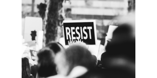 Black and white photo of an outdoor protest with one sign that says RESIST in white letters against a black piece of cardboard