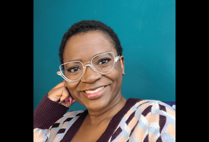 Graphic with a black background with a photo of a Black female with dark brown complexion, close cropped black hair, wearing a wide smile, clear plastic glass frames and sweater with a geometric pattern in eggplant, sky blue, white and light peach colors.