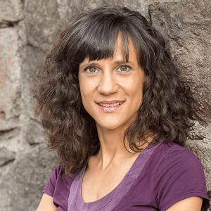 Headshot of a white woman with shoulder-length curly brown hair. She leans against a rough rock wall and smiles.