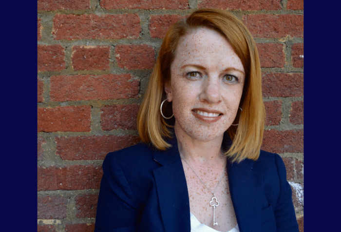 Graphic with a navy blue background and in the center a photo of a red-haired and freckled woman wearing a blue blazer and standing in front of a red brick wall
