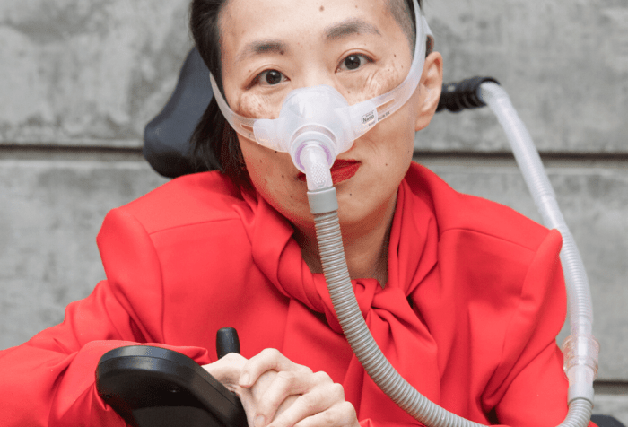 Photo of an Asian American woman in a power chair. She is wearing an orange-red jacket and black pants. She is wearing a mask over her nose attached to a gray tube and bright red lip color. Her hands are resting over her joystick. Photo credit: Eddie Hernandez Photography