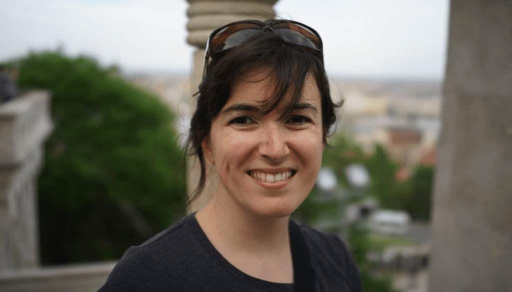 Image Description: A light-skinned woman of Brazilian heritage is standing outside and smiling at the camera. Her eyes twinkle at you, framed by dark and bold eyebrows. Her dark brown hair is pushed back by a pair of sunglasses, tendrils of hair fanning out around her face. Tactile Description: Cristina Hartmann has small, smooth hands that move deliberately and precisely, often punctuating her thoughts with a wry pinky wriggle-laugh. Her most distinctive tactile feature is her hair, soft and coconut-scented, which she keeps pulled back into a twist. She leans comfortably to the side as she chats with someone, always a bit askew.
