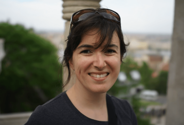 Image Description: A light-skinned woman of Brazilian heritage is standing outside and smiling at the camera. Her eyes twinkle at you, framed by dark and bold eyebrows. Her dark brown hair is pushed back by a pair of sunglasses, tendrils of hair fanning out around her face. Tactile Description: Cristina Hartmann has small, smooth hands that move deliberately and precisely, often punctuating her thoughts with a wry pinky wriggle-laugh. Her most distinctive tactile feature is her hair, soft and coconut-scented, which she keeps pulled back into a twist. She leans comfortably to the side as she chats with someone, always a bit askew.