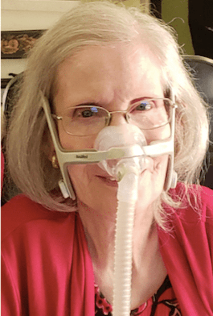 Headshot image of Diane Coleman wearing red print top and red sweater, smiling with gray bobbed hair, wire rimmed glasses and a nasal breathing mask with a white tube hanging downward.