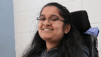 A South Asian woman with light brown skin, dark eyes, and black glasses smiles at the camera. She is sitting in an electric wheelchair with a purple jacket tucked behind the headrest, her hands folded over a Chromebook that rests on a wheelchair tray. Behind her is a white brick wall that gives way to untextured blue on the left of the image.