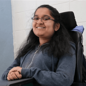 A South Asian woman with light brown skin, dark eyes, and black glasses smiles at the camera. She is sitting in an electric wheelchair with a purple jacket tucked behind the headrest, her hands folded over a Chromebook that rests on a wheelchair tray. Behind her is a white brick wall that gives way to untextured blue on the left of the image.