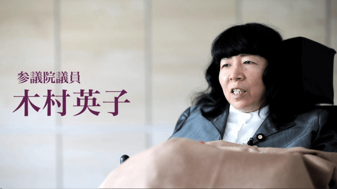 Graphic showing a picture of Eiko Kimura, a Japanese woman in a wheelchair. She has long black hair and wearing a navy jacket and a white shirt. A beige blanket is on her lap. On the right in Japanese 参議院議員 木村英子is text translated: Upper House Member (or member of the House of Councillors), Kimura Eiko.