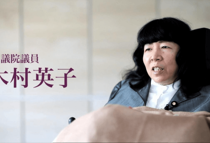 Graphic showing a picture of Eiko Kimura, a Japanese woman in a wheelchair. She has long black hair and wearing a navy jacket and a white shirt. A beige blanket is on her lap. On the right in Japanese 参議院議員 木村英子is text translated: Upper House Member (or member of the House of Councillors), Kimura Eiko.