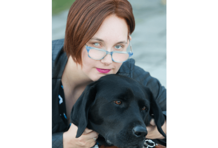 Elsa, a caucasian woman with short asymmetrical red hair, looks over her blue glasses with a half smile on her face. Her right eye is an aqua cataract, her right is grey and has a pupil. She wears pink lipstick that matches her pink and grey hearing aids. She wears a teal geometric blouse and leather jacket, and is pictured with her guide dog. Her guide dog has brown eyes and looks bored.