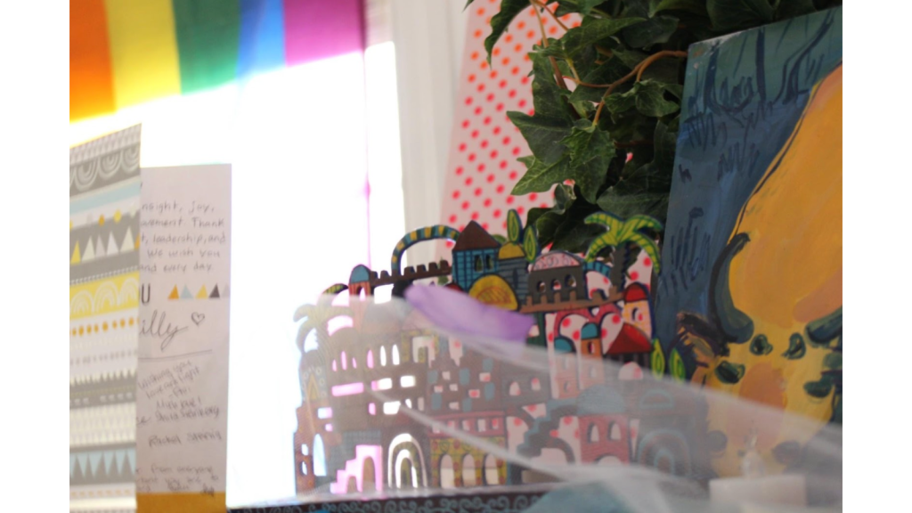 [A multicolored ceramic portrayal of the Jerusalem skyline sits on a shelf. A card is next to it. In the background there is a rainbow pride flag.]