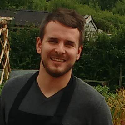 Photo of a white man with short brown hair and beard. He is wearing a dark gray v-neck t-shirt and black apron. Behind him are trees and a house.