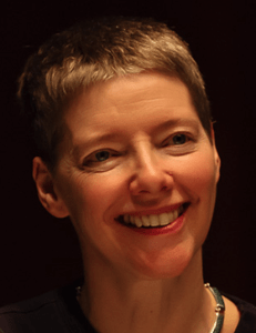 Photo of a white woman with short blonde hair. She is smiling and looking partially sideways from the camera. The background behind her is completely black. She has a black scoop neck shirt and a necklace made of a silver or silver-looking metal.