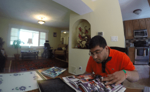 Young Pakistani American man with dark hair and glasses sitting at a table reading a comic book.