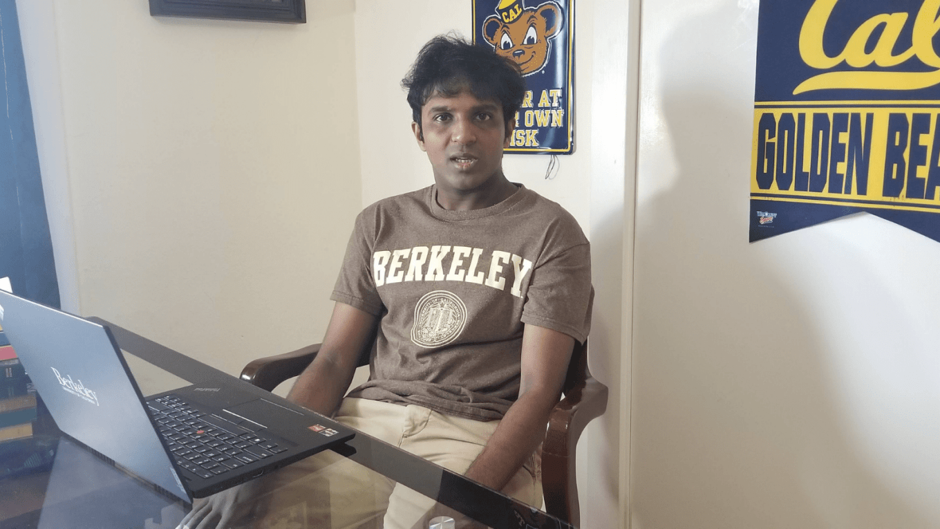 The image shows a young American male in his 20s of Indian American descent, with brown skin and black hair. He is sitting at a desk with a laptop open in front of him with some books on the side of the table. The laptop has the word Berkeley on it as does the brown t-shirt he is wearing. Behind him on the wall are college banners and posters with words like Cal, Golden Bears, and catchy phrases like, Bear Den, enter at your own risk.