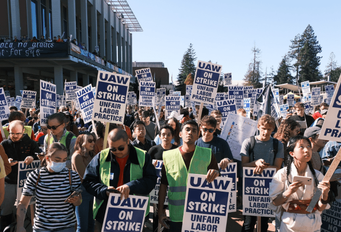 Many, many workers with picket signs reading "UAW: On Strike: Unfair Labor Practice". They are crowded together outdoors and in the sea of faces there appears to be only one worker wearing a surgical mask. Photo by: Ian Castro. 2022. Provided on FairUC Now Website for Press Use