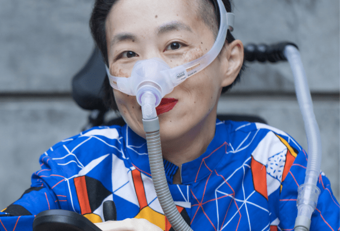 Photo of an Asian American woman in a power chair. She is wearing a blue shirt with a geometric pattern with orange, black, white, and yellow lines and cubes. She is wearing a mask over her nose attached to a gray tube and bright red lip color. She is smiling at the camera. Photo credit: Eddie Hernandez Photography