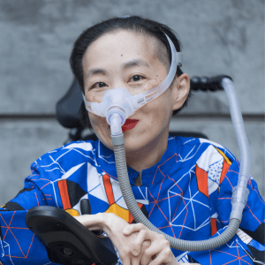 Photo of an Asian American woman in a power chair. She is wearing a blue shirt with a geometric pattern with orange, black, white, and yellow lines and cubes. She is wearing a mask over her nose attached to a gray tube and bright red lip color. She is smiling at the camera. Photo credit: Eddie Hernandez Photography