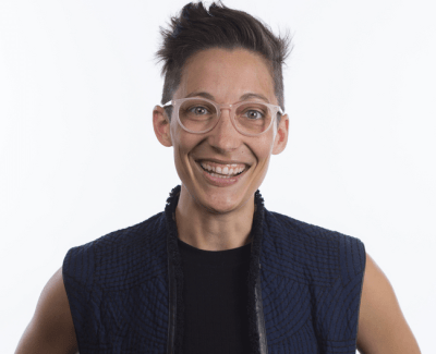 Photo of Liz Jackson, a queer white disabled person with short hair. She is wearing eyeglasses, a navy vest with a black t-shirt underneath. She is smiling broadly. Photo credit: Ryan Lash