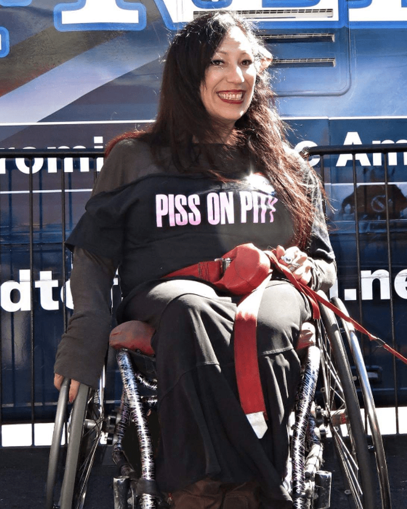 Una foto de María, una mujer latina de pelo largo y oscuro que lleva una camiseta que dice "Piss On Pity", con los labios rojos y una enorme sonrisa. Está sentada en su silla de ruedas delante de una valla con un gran cartel o vehículo detrás de ella. Tiene una correa roja alrededor de la cintura, conectada a algo fuera de la pantalla. Parece estar en una protesta o acción directa