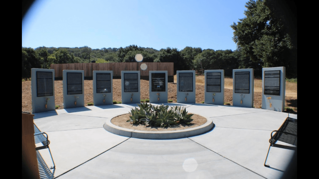 Graphic with a black background with a photo of a monument on the grounds of Napa State Hospital in Northern California. It consists of several gray pillars with black displays with white texts, too far away to read. In the foreground are several large spiky succulent plants. In the background are a wooden fence and trees underneath a blue sky.