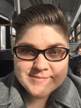 Photo shows a close up image of a Kate, a not-as-young-as-she-used-to-be white person wearing thick rimmed glasses. She is smiling while sitting on Chicago public transit in her grey pea coat. She has short cropped brown hair that is trimmed close on the sides, but longer on top and swept over and back to one side.