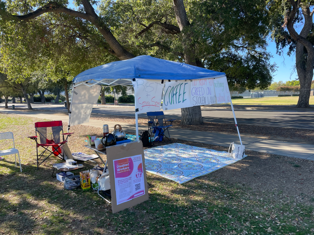 The Mutual Aid/Disability Tent at UC Davis is a tent under an oak tree. It is set up next to a walkway on the flatter, more smooth area of the lawn where the strike line was set up. Beneath it are blankets and chairs, food and water. The signs around it read "Rest + Sensory Space" "Coffee" "No Room for Greed on Stolen Land" and "Nothing About Us Without Us" It also includes an informational sign about Disabled Workers and the Strike (the detailed text illegible in this photograph). Photo of UC Davis Mutual Aid Tent by Breanne Weber. 2022. Used with permission.
