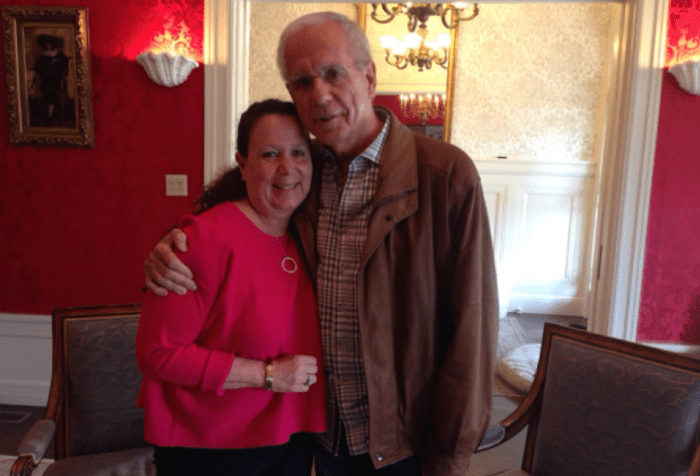 Image of two people standing next to each other. On the left is an older white woman with long brown curly hair. She is wearing a red sweater. Next to her is a tall older Hispanic man wearing a brown leather jacket. He has white hair and eyeglasses. His hand is around her shoulder.
