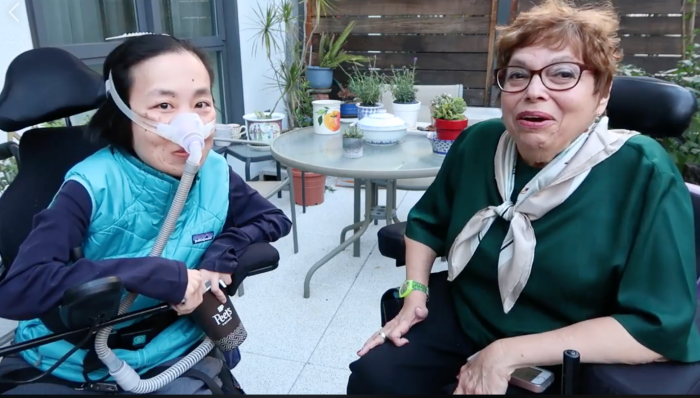 An outdoor patio with Alice Wong [left], an Asian American disabled woman in a wheelchair. She is wearing a mask over her nose connected to a tubing for her ventilator. She is wearing a long-sleeved navy shirt and a sporty aqua blue vest and holding a paper coffee cup. On the right is Judith Heumann, an older white woman with short wavy brown hair. She is sitting in a power chai and wearing glasses, a dark green short-sleeved top and a scarf tied into a knot. Both are looking at the camera and smiling. 
