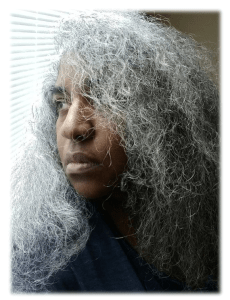 Image of an Afro-Latina woman with long curly gray hair. She is staring out at the window that has white blinds. 