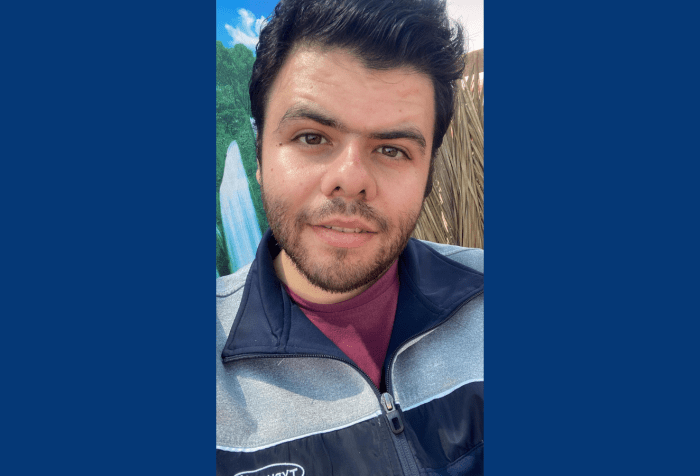 Graphic with a dark blue background. In the center is a close-up of Khaled outside in the sun, dark brown hair combed up, slight brown beard and hazel-brown eyes staring into the camera. He wears a slight smile and a blue and gray zip up with maroon shirt underneath. 
