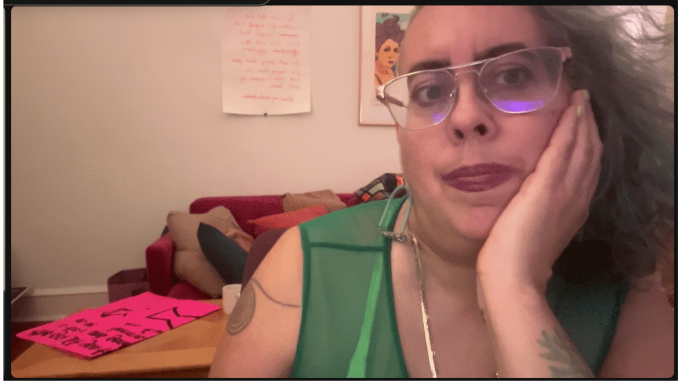 Leah, a nonbinary Sri Lankan and white femme in their 40s with sand colored skin, grey smoke, chestnut brown and faded teal curls on one side of their head and a shaved side on the other, rose gold glasses and purple lips, looks at the viewer with their head resting on their left hand. They wear a apple green mesh top through which a neon yellow bra strap is visible. They sit in front of a hot pink couch heaped with cushions and a blonde wood coffee table with a pink protest sigh on it.