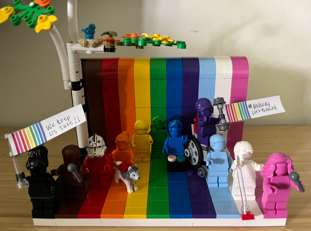 This is a Lego rainbow Everyone is Awesome set with upgrades. It includes the rainbow minifigures and some have mobility devices and some are holding protest signs. In the background is a Lego tree with a blue bird in it and orange and yellow leaves. The Black minifigure is holding a sign that says “We Keep Us Safe.” The brown mini figure is holding a microphone. The red one is wearing a storm trooper mask. The orange one is sitting down with a service animal dog. The yellow one has a limb difference. The blue one has an AAC device in front of it. The purple one is in a wheelchair holding a protest sign “# No Body Left Behind. The white one is holding a guide cane. And the pink one is holding a microphone.