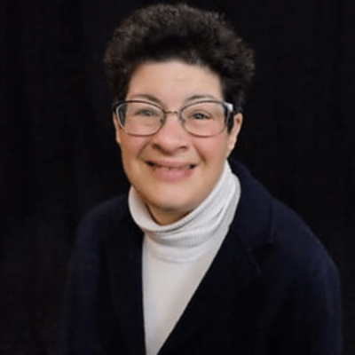 A picture of a woman with a blue sweater and a white turtleneck with glasses and smiling for the camera.