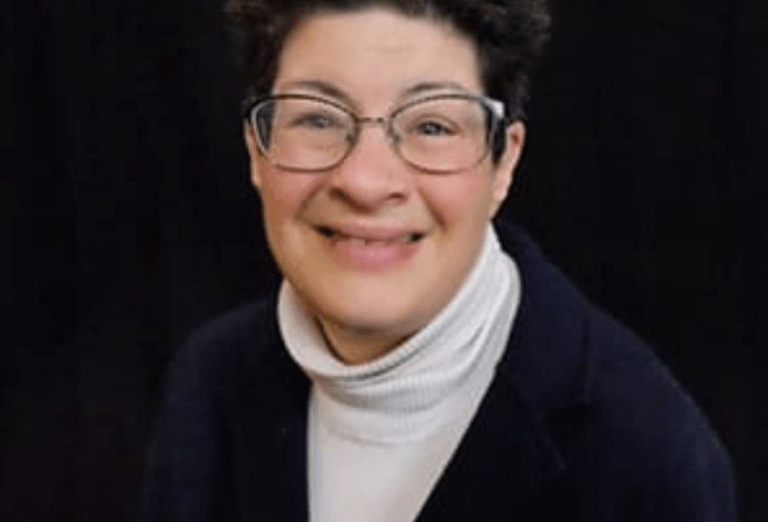 A picture of a woman with a blue sweater and a white turtleneck with glasses and smiling for the camera.